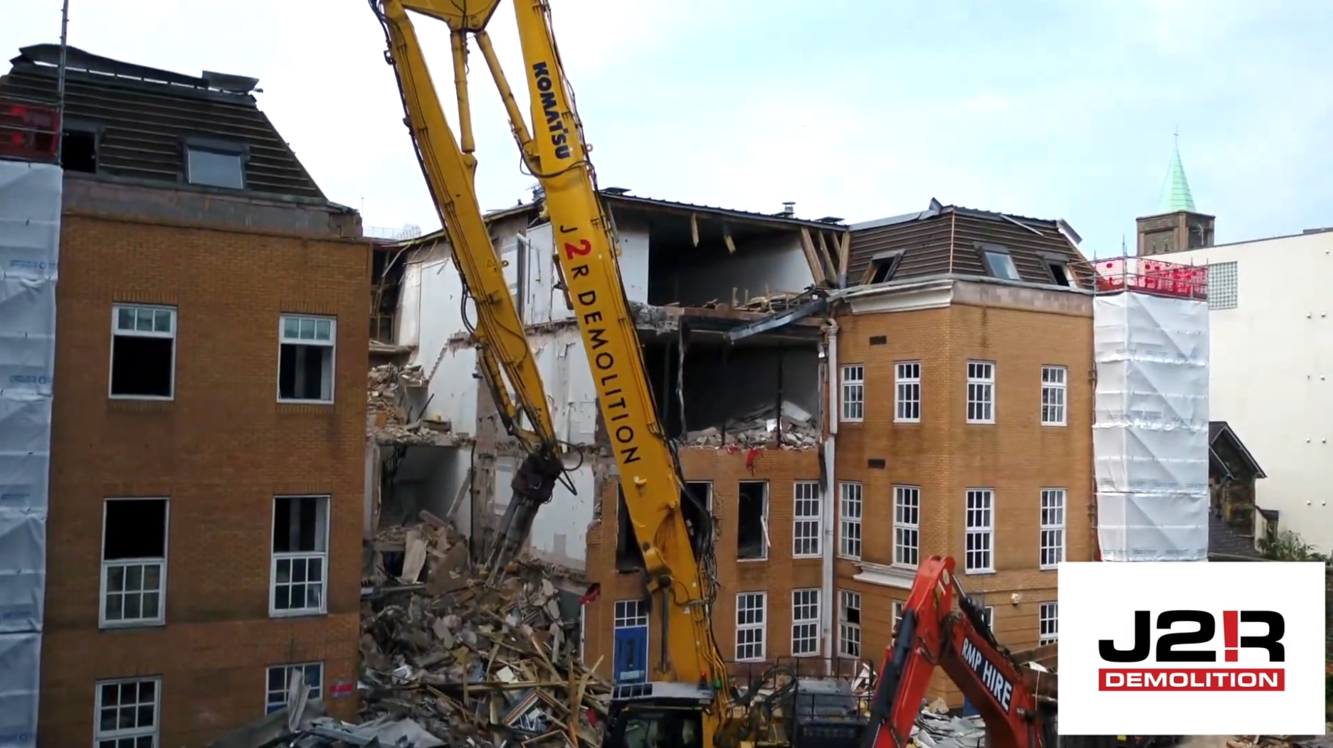 Excavator demolishing a multi-storey brick building, with rubble in the foreground and J2R Demolition branding visible.